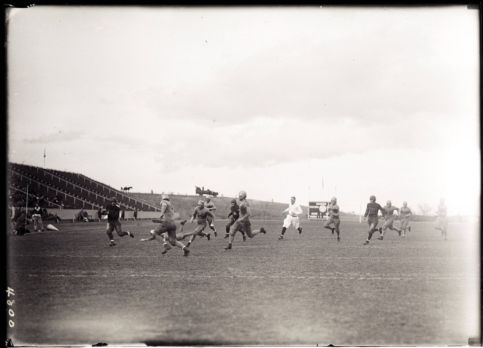 Thirteen players run across the field toward player in dark uniform carrying the ball; a horse and a car are visible on a hill in the background.