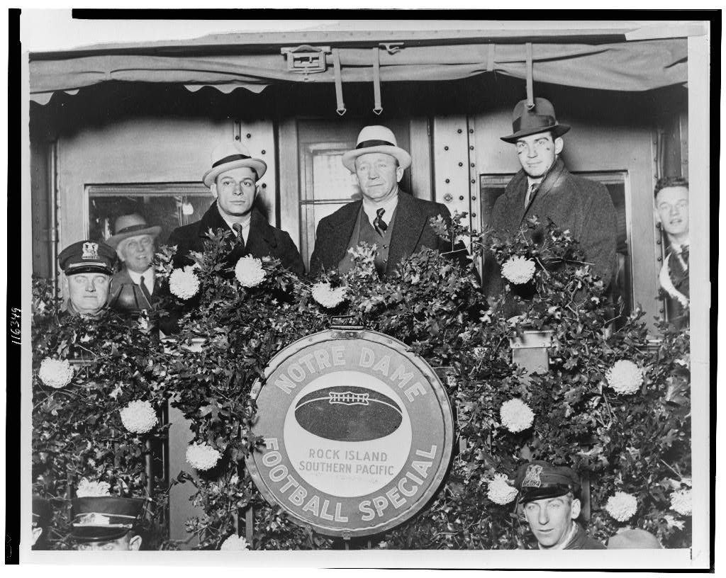Knute Rockne standing outside rear of train car, flanked by Mr. Cariedo and Mr. Conley, members of the Notre Dame football team, before departing to California